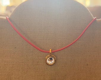 Può includere: Una collana con cordino rosso con un ciondolo color oro. Il ciondolo presenta una gemma rotonda e trasparente incastonata in una lunetta color oro. La collana è esposta su una superficie di colore neutro.