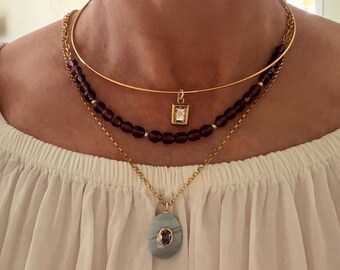 May include: A layered necklace set featuring three distinct pieces. The top layer is a gold choker with a small square pendant. The middle layer has dark red beads. The bottom layer has a gold chain with a blue-grey oval pendant.