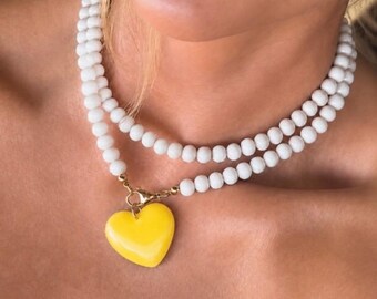 May include: A double-strand necklace featuring small, round, opaque white beads. The necklace is secured with a gold-toned metal lobster clasp that holds a single, smooth, bright yellow heart-shaped pendant hanging at the center of the collar.