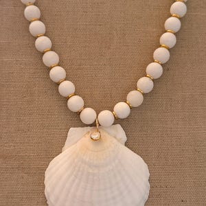 May include: A white beaded necklace with a large white seashell pendant. The necklace features round white beads separated by gold-colored spacers. A small, round, clear gemstone hangs from the necklace, attached to the seashell.