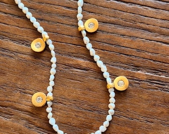 May include: Delicate necklace featuring a strand of small, white pearls. Gold-colored circular charms with a central clear stone are evenly spaced along the necklace. The necklace is displayed on a wooden surface.