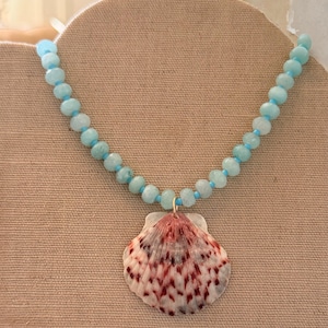 May include: A necklace featuring a large seashell pendant with red and white patterns, suspended from a strand of light blue beads. The necklace is displayed on a neutral-toned surface.