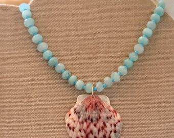 Può includere: Una collana con un ciondolo a conchiglia con motivi rossi e bianchi, sospeso a una fila di perline azzurre. La collana è esposta su una superficie dai toni neutri.