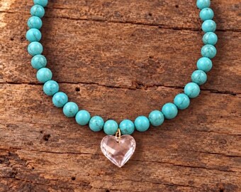May include: A necklace featuring a strand of round, bright turquoise beads with a clear, faceted, heart-shaped pendant suspended from a small gold-toned metal jump ring at the center of the beaded strand.
