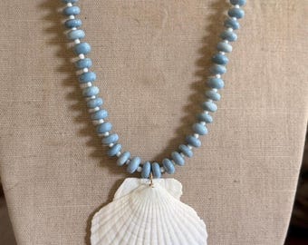 Può includere: Una collana con un grande ciondolo a forma di conchiglia bianca a ventaglio, sospeso a un filo di perline azzurre a forma di disco. Le perline sono separate da piccoli distanziatori sferici bianchi, creando un motivo ripetitivo lungo l'intera lunghezza della catena.