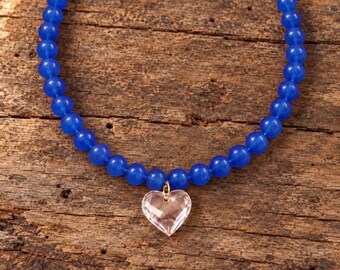 May include: A necklace featuring a strand of round, translucent blue beads. A single, faceted, clear heart-shaped pendant is attached to the center of the bead strand with a small gold-toned metal jump ring, creating a simple and symmetrical design.
