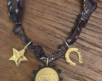 May include: A black and white paisley print fabric bandana necklace featuring three gold-toned metal charms. The charms include a five-pointed star, a horseshoe, and a circular pendant with a raised horse figure surrounded by small black spheres.