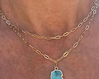 May include: A gold-toned, layered necklace with a rectangular turquoise pendant. The pendant has a silver-colored border. The necklace is made of linked chain. The necklace is displayed on a person's neck.