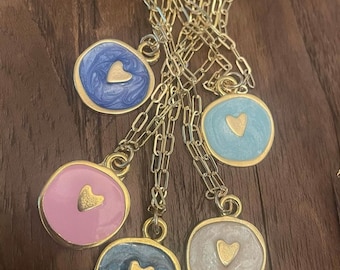 Può includere: Cinque collane d'oro con ciondoli rotondi in resina di diversi colori e un ciondolo a forma di cuore in oro. I ciondoli sono blu, azzurro, rosa, bianco e grigio.