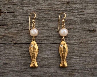 Può includere: Un paio di orecchini pendenti color oro con un ciondolo a forma di pesce dalla texture martellata e un piccolo cristallo trasparente. Ogni orecchino è sospeso a un gancio dorato e include una perla bianca rotonda sopra il ciondolo a pesce.