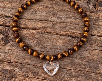 May include: A beaded necklace featuring round, polished tiger's eye gemstones in shades of brown and gold. The necklace is strung on a cord and includes a single, clear, faceted heart-shaped pendant suspended from a small gold-toned metal jump ring.