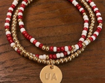 Può includere: Un set di tre braccialetti sovrapposti con perline rosse e bianche alternate, perline dorate metallizzate e un ciondolo circolare color oro inciso con le lettere UA. I braccialetti sono realizzati con un cordino elastico flessibile.