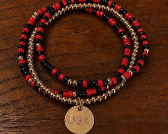 Può includere: Un bracciale a più fili di perline che alterna perline rosse, nere e oro metallizzato. Il bracciale include un ciondolo circolare in metallo dorato inciso con le lettere UGA in un semplice carattere sans-serif, centrato nella parte inferiore.