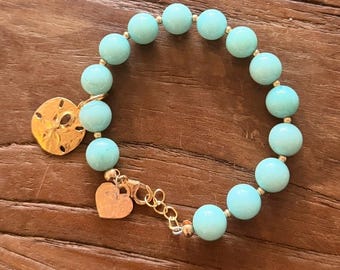 Può includere: Un braccialetto di perline azzurre con dettagli dorati. Include un ciondolo a forma di dollaro di sabbia dorato e un ciondolo a forma di cuore. Il braccialetto è su una superficie di legno.
