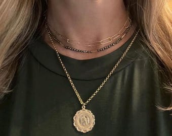 Può includere: Un set di collane a strati con tre catene dorate. La catena superiore è un delicato design a maglie, seguito da una catena di perline e una catena più lunga con un ciondolo circolare. Il ciondolo ha un design in rilievo.