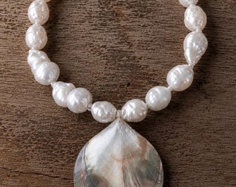 Può includere: Una collana composta da un filo di perle barocche bianche di forma irregolare. La collana è completata da un grande ciondolo a forma di goccia in conchiglia iridescente, che presenta sfumature di grigio, argento e delicati toni rosati.