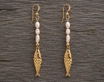 Può includere: Un paio di orecchini pendenti color oro con un ciondolo a forma di pesce alla base. Ogni orecchino è sospeso a un gancio curvo e presenta una disposizione verticale di tre perle bianche ovali posizionate sopra il ciondolo a forma di pesce.
