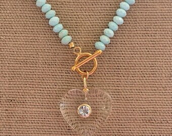 May include: A necklace featuring light blue beaded strands and a gold-colored clasp. A clear heart-shaped pendant with a small, round, light blue stone is suspended from the clasp. The necklace is displayed on a textured, tan surface.