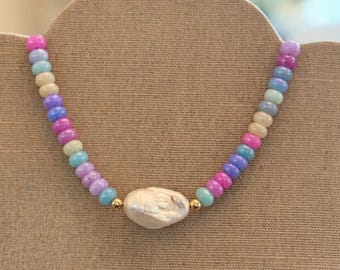 Può includere: Una collana di perline colorate con un grande pendente di perla bianca irregolare. Le perline sono in tonalità pastello rosa, blu, giallo e viola. Perline color oro su entrambi i lati della perla. La collana è esposta su un espositore per gioielli dai toni neutri.