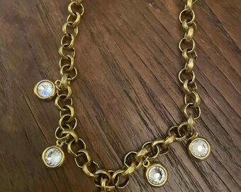 May include: Gold-toned necklace with a chunky chain design. The necklace features several round, clear crystal pendants. The crystals are set in gold-colored bezels. The necklace is displayed on a dark wooden surface.