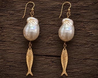 Può includere: Un paio di orecchini pendenti con grandi perle barocche bianche irregolari sospese a ciondoli dorati a forma di pesce. Gli orecchini sono fissati a ganci dorati, mostrando una combinazione di texture di perle organiche e motivi di pesci metallici.