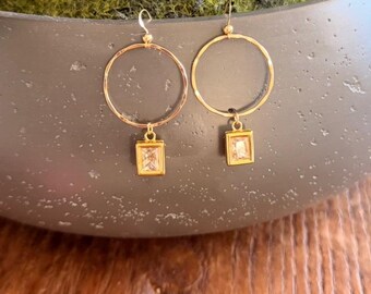 Può includere: Un paio di orecchini a cerchio color oro. Ogni orecchino presenta un grande cerchio circolare con un piccolo pendente quadrato. I pendenti hanno una pietra rettangolare trasparente incastonata in una cornice dorata. Gli orecchini sono su sfondo neutro.