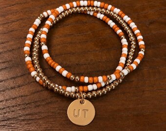 Può includere: Un set di tre braccialetti sovrapposti con perline arancioni e bianche alternate, perline dorate metallizzate e un ciondolo circolare color oro inciso con le lettere UT, disposti in un motivo circolare su una superficie.