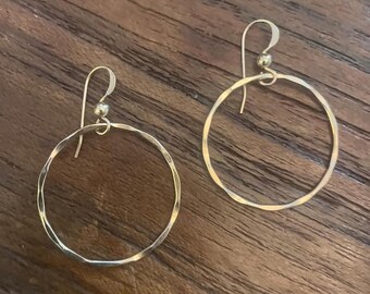 May include: A pair of gold-colored hoop earrings with a hammered texture. The earrings have a simple, elegant design with a circular shape and a hook closure. The earrings are displayed on a wooden surface.