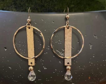 Può includere: Un paio di orecchini dorati. Ogni orecchino presenta una barra dorata martellata sospesa all'interno di un cerchio dorato strutturato. Una piccola perla dorata e un cristallo trasparente a forma di goccia pendono in basso.