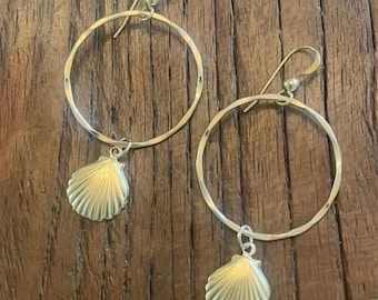 Può includere: Un paio di orecchini a cerchio color argento con una sottile struttura in filo circolare e un ciondolo a forma di conchiglia. Ogni orecchino è dotato di un gancio curvo per l'attacco, con una finitura metallica dal riflesso sottile.