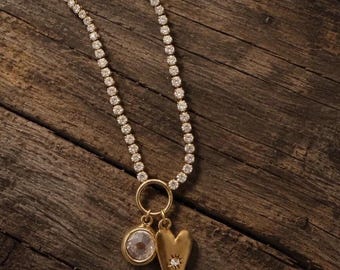 Può includere: Una collana tennis color oro con una fila continua di pietre trasparenti a taglio rotondo. La collana presenta un anello pendente che sostiene due ciondoli: una pietra rotonda incastonata e un cuore dorato con una piccola pietra a stella.