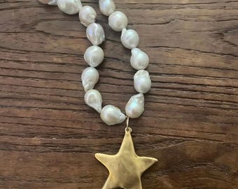 Può includere: Una collana di perle bianche con un ciondolo a forma di stella dorata. La collana presenta grandi perle di forma irregolare infilate insieme. Il ciondolo a forma di stella dorata pende dal centro della collana. La collana è esposta su una superficie di legno.