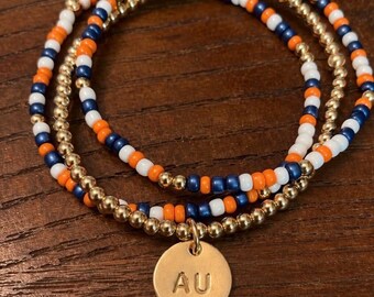 Può includere: Un set di tre braccialetti sovrapposti con perline alternate arancioni, bianche e blu scuro, intervallate da perline color oro metallizzato. Un braccialetto include un ciondolo rotondo e piatto color oro con incise le lettere AU.