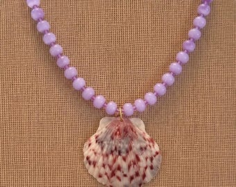 Può includere: Una collana con un ciondolo a forma di conchiglia con motivi rossi e bianchi, sospeso a una fila di perline viola chiaro. La collana è esposta su uno sfondo neutro, evidenziando i colori e il design del gioiello.