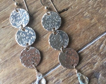 Può includere: Un paio di orecchini dorati con tre dischi d'argento martellati e un cristallo trasparente a forma di lacrima che pende da ogni orecchino.
