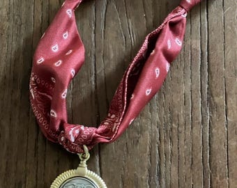 May include: A burgundy bandana necklace with a gold-colored pendant. The pendant features an elephant design within a circular frame. The bandana has a paisley pattern and is knotted at the top.