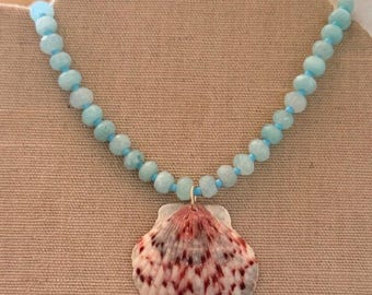 Può includere: Una collana composta da una fila di perline sfaccettate azzurre alternate a piccole perline distanziatrici blu brillante. Un singolo grande ciondolo a forma di conchiglia a ventaglio con base bianca e motivi maculati rosso-bruni.
