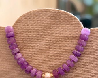 Può includere: Una collana di pietre preziose viola con un accento di perla. La collana presenta perline viola rettangolari e rotonde, con una singola perla bianca e una perlina color oro al centro. Viene esposta su un tessuto beige.