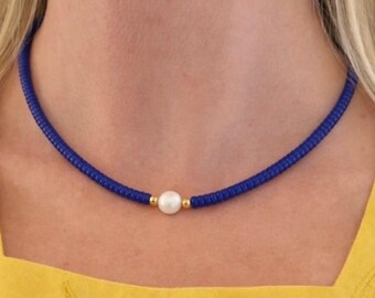 Può includere: Una collana caratterizzata da un filo continuo di piccole perle cilindriche color blu reale. Una singola perla sferica bianca è posizionata al centro, affiancata da due piccole perle metalliche color oro su entrambi i lati.