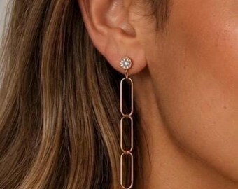 May include: A rose gold drop earring featuring a small circular stud set with a clear stone, connected to a vertical chain of three elongated oval links. The earring has a polished metallic finish and hangs in a straight line from the earlobe.