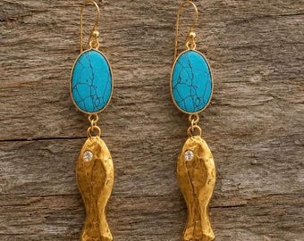 Può includere: Un paio di orecchini pendenti color oro con una pietra turchese ovale con venature scure nella parte superiore, collegata a un ciondolo dorato testurizzato a forma di pesce con un piccolo cristallo trasparente vicino alla parte superiore.