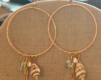 May include: Gold hoop earrings with seashell, chain, and crystal charms. The earrings have a hammered texture and are displayed on a neutral background. The seashell charms have brown and white stripes.