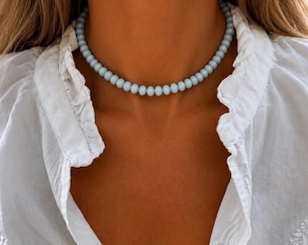 Può includere: Una collana girocollo di perline azzurre composta da un unico filo di perline uniformi, rotonde e lisce. La collana aderisce al collo, creando un contrasto con una camicia bianca con colletto e bordo arricciato lungo l'abbottonatura.