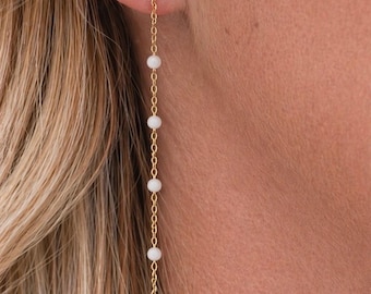 Può includere: Orecchini pendenti color oro con un piccolo perno a sfera e una delicata catena. La catena sospende una serie di piccole perle rotonde bianche. Gli orecchini sono mostrati su uno sfondo di capelli biondi e pelle.