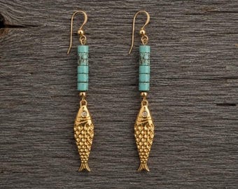 Può includere: Un paio di orecchini pendenti color oro con un ciondolo a forma di pesce dotato di una piccola pietra chiara sulla testa. Sopra il pesce, quattro perline cilindriche color turchese sono impilate verticalmente, sospese a un gancio dorato.