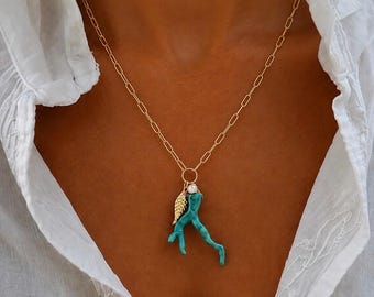 Può includere: Una collana a catena color oro con un ciondolo composto da un elemento in corallo turchese a forma di ramo, un piccolo ciondolo a forma di ala dorata testurizzata e una singola perla bianca, sospesi da un anello di giunzione circolare dorato.