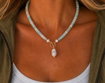 May include: A beaded necklace featuring light blue and white round beads, accented with a gold-toned toggle clasp at the center. A single, irregular white baroque pearl pendant hangs from the gold toggle ring, creating a focal point.