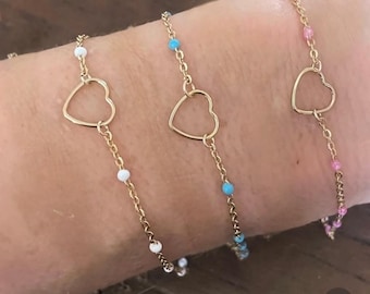 Può includere: Tre braccialetti a catena dorati con ciondoli a forma di cuore. Ogni braccialetto presenta piccole perline di diversi colori: bianco, turchese e rosa. I braccialetti sono mostrati su un braccio.