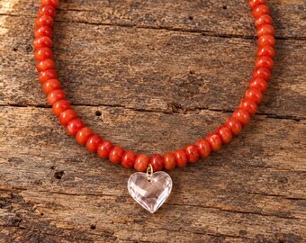May include: A necklace featuring a strand of small, round, vibrant orange-red beads. A single, clear, faceted heart-shaped pendant hangs from the center of the bead strand, attached by a small gold-toned metal jump ring.