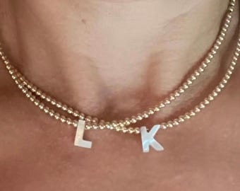 Può includere: Collane dorate con perline con le lettere "L" e "K" come ciondoli. Le collane sono a strati e poggiano sul collo di una persona. Le lettere sono bianche e hanno un aspetto madreperlaceo.